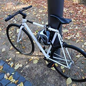 Ein weißes Fahrrad, das an einem Pfahl mit der Aufschrift Langes Kettenschloss mit Zahlenkombination "Oslo" und einem Helm befestigt ist, inmitten von trockenem Gras und herabgefallenen Blättern in Oslo.