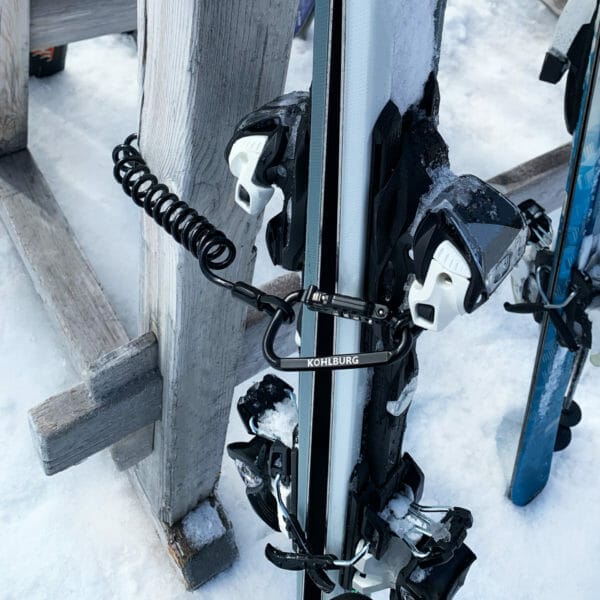 Nahaufnahme des Premium Kabelschlosses mit Zahlenkombination "Zermatt", das ein Paar Skier auf einem Holzgestell auf verschneitem Boden sichert.