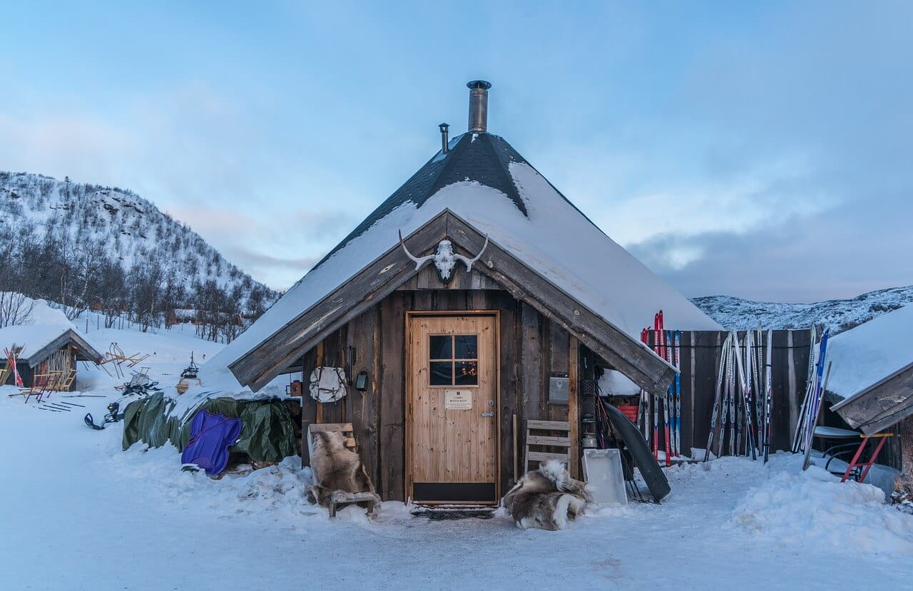 Verschneite Holzhütte mit Skiern und Fellteppichen im Freien in einer verschneiten Berglandschaft.