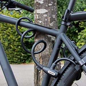 Das Langes Kabelschloss mit Zuklick-Mechanismus "Paris" sichert ein schwarzes Fahrrad an einem Baum. Das Foto zeigt die Szene im Freien, umgeben von viel Grün.