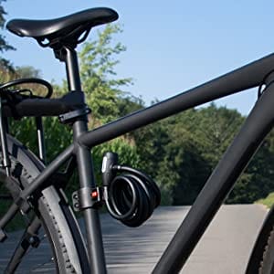 Schwarzes Fahrrad mit einem am Rahmen befestigten Langes Kabelschloss mit Zuklick-Mechanismus "Paris", geparkt auf einem gepflasterten Weg mit Bäumen im Hintergrund.