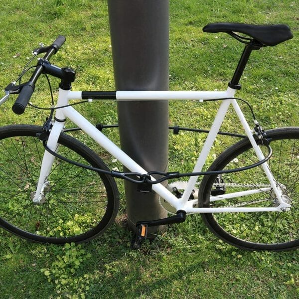 Ein weißes Fahrrad ist mit dem "Athen"-Kabelschloss - einem robusten Stahlkabel und Vorhängeschloss - an einem Pfosten im Gras befestigt.