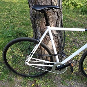 Ein weißes Fahrrad ist an einem Baum mit dem Kabelschloss "Athen" - Stahlseil & Vorhängeschloss befestigt und auf einer von grünem Laub umgebenen Wiese abgestellt.