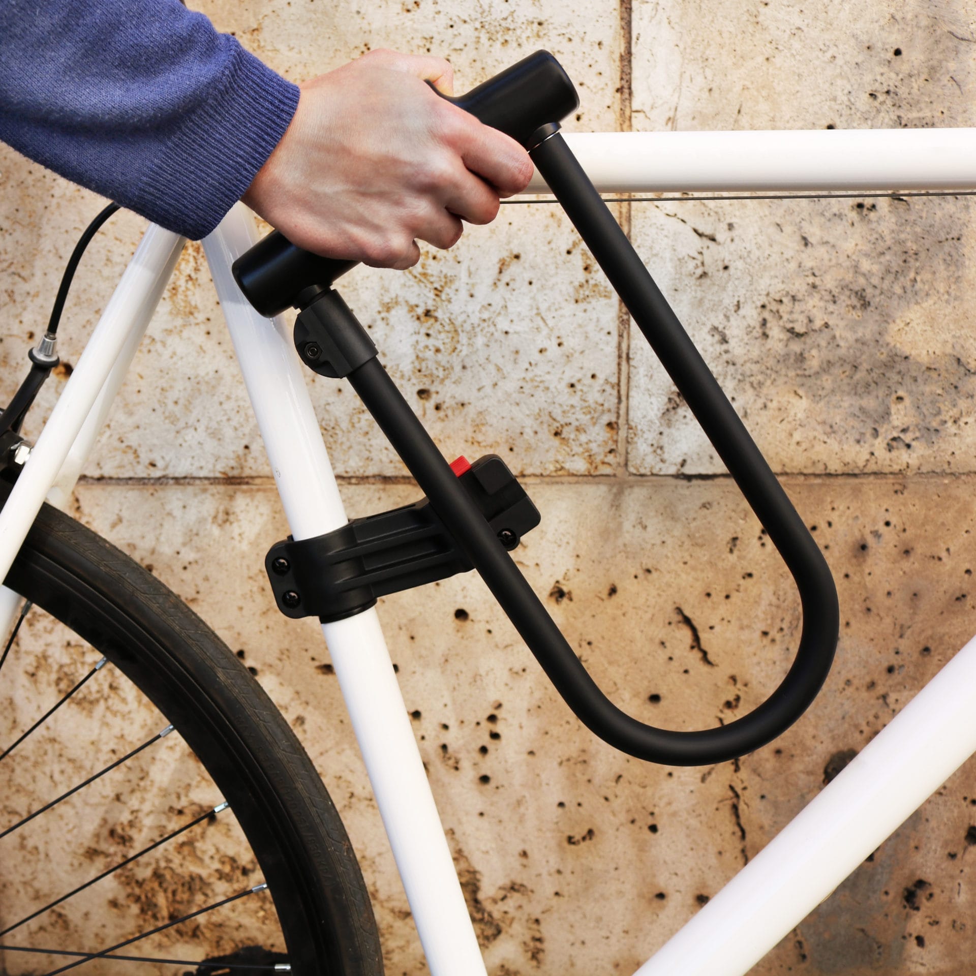 Eine Person sichert einen weißen Fahrradrahmen an einer Steinmauer mit dem Sicherheits-Bügelschloss "Novosibirsk" inkl. Sicherheitskabel, einem schwarzen Bügelschloss.