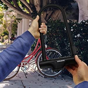 Person hält das Sicherheits-Bügelschloss "Novosibirsk" incl. Sicherheitskabel vor einem roten Fahrrad, das an einem Baum auf sonnigem Bürgersteig steht.