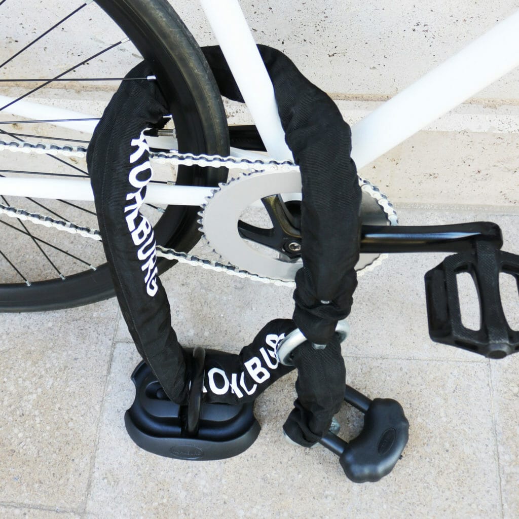 Ein Fahrrad wird mit dem Sicherheits-Bodenanker "London", einem Kettenschloss, das um den Rahmen und das Hinterrad gewickelt wird, an einem Fahrradständer befestigt.