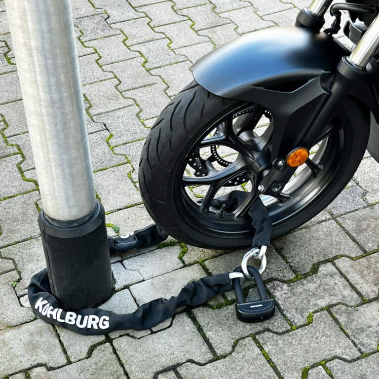 Das Vorderrad eines Motorrads wird mit einem Schloss und einer Kette auf einer gepflasterten Fläche an einer Metallstange befestigt.