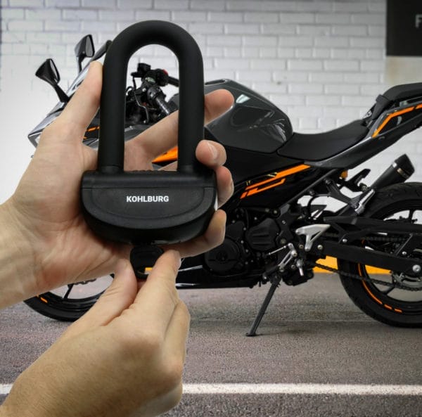 Eine Person hält ein KOHLBURG-Schloss vor ein schwarzes Motorrad, das an einer weißen Backsteinmauer geparkt ist.