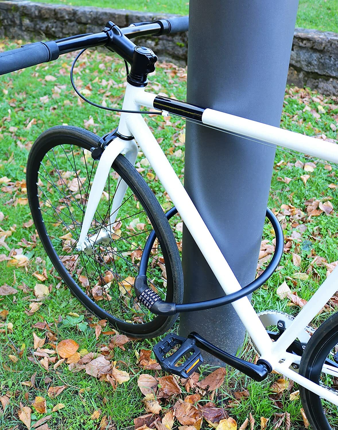 Das lange, massive Kabelschloss mit Zahlenkombination "Bordeaux" sichert ein weißes Fahrrad am Rad statt am Rahmen an einem Pfosten, geparkt auf Gras mit Herbstlaub.