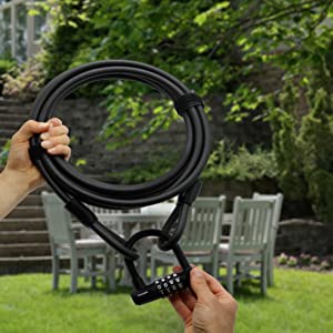 Person, die das Kabelschloss "Sparta" - Stahlseil & Zahlenschloss im Freien neben einem Terrassentisch und Stühlen in einem Garten hält.