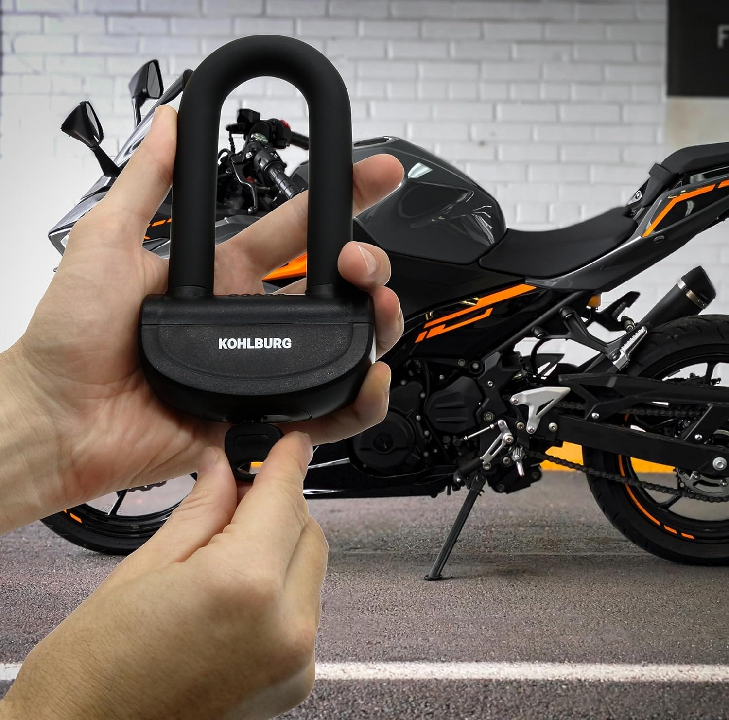 Hände, die ein Sicheres Motorradschloss "Prag" mit einem Schlüssel halten, neben einem schwarz-orangen Motorrad vor einer Backsteinmauer.