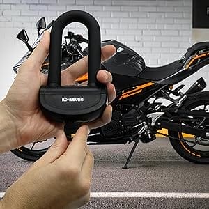 Hände halten das schwarze Sichere Motorradschloss "Prag" vor einem geparkten Motorrad mit orangenen Akzenten.