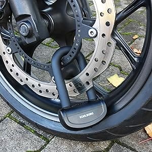 Nahaufnahme des Sicheres Motorradschloss "Prag", ein schwarzes Bügelschloss, das ein Motorradrad und eine Bremsscheibe auf einem Steinpflaster sichert.